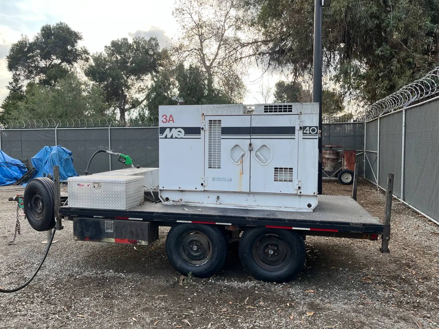 Trailer-mounted generator with a fuel tank and green dispensing nozzle parked on a gravel lot