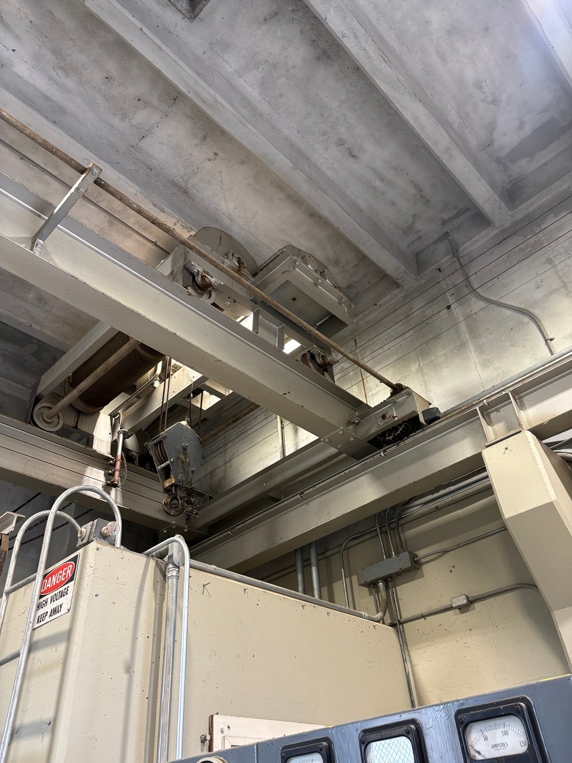 Overhead bridge crane with cable drum and suspended hook above a beige high-voltage enclosure marked DANGER HIGH VOLTAGE KEEP AWAY, inside a concrete industrial hall