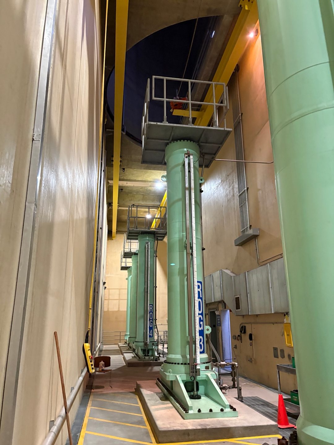 Row of tall pale-green vertical operating cylinders labeled ROG 3 and ROG 4 bolted to concrete plinths inside an indoor equipment gallery with an overhead yellow crane rail